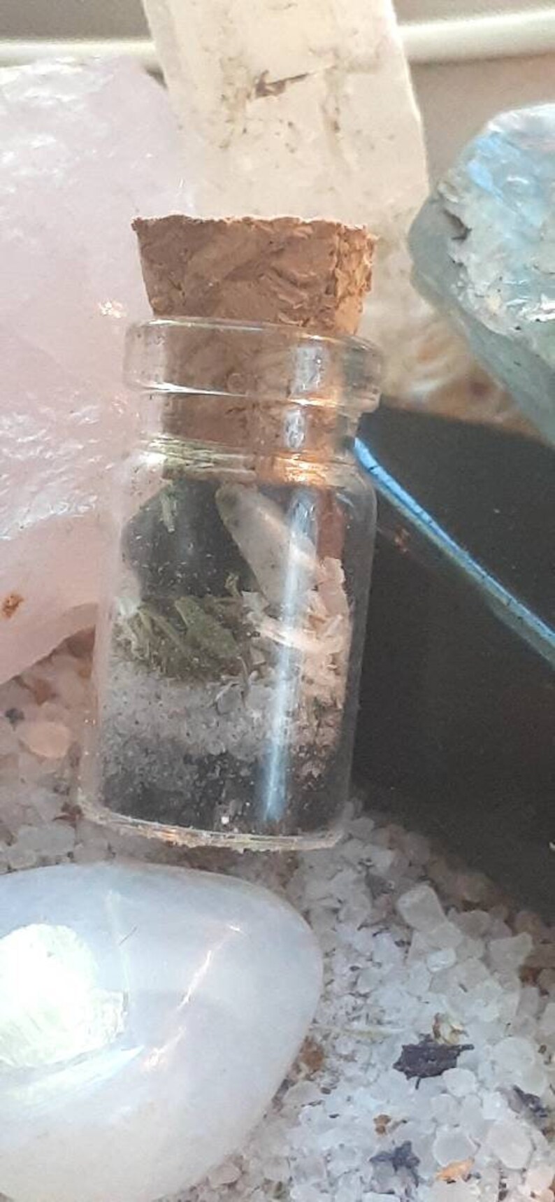 May include: A small glass bottle with a cork stopper, filled with dried herbs and crystals. The bottle is sitting on a white surface with other crystals.