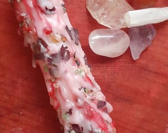 May include: A pink and white candle with red and green herbs embedded in the wax. The candle is surrounded by clear quartz, rose quartz, and carnelian crystals.