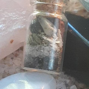 May include: A small glass bottle with a cork stopper filled with white and green crystals. The bottle is sitting on a white surface.