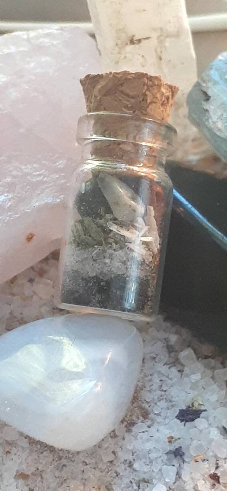 May include: A small glass bottle with a cork stopper filled with white and green crystals. The bottle is sitting on a white surface.