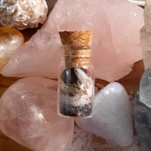 May include: A small glass bottle with a cork stopper filled with various crystals, herbs, and other natural elements. The bottle is sitting on a pink and white surface.