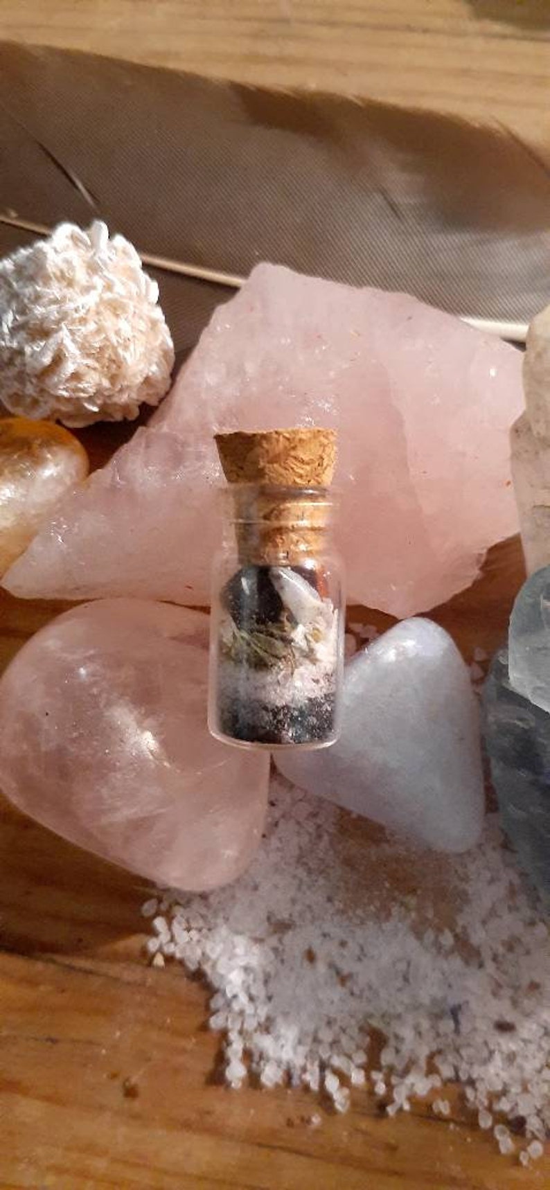 May include: A small glass bottle with a cork stopper filled with various crystals, herbs, and other natural elements. The bottle is sitting on a pink and white surface.