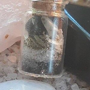 May include: A small glass bottle with a cork stopper, filled with dried herbs, crystals, and a small white feather. The bottle is sitting on a white surface.