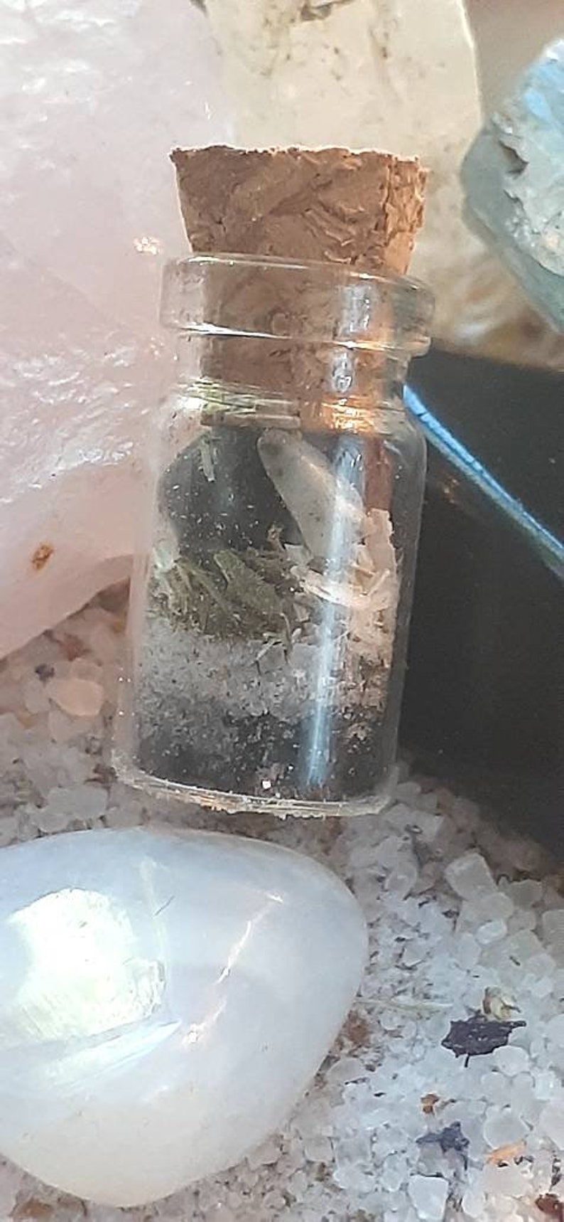 May include: A small glass bottle with a cork stopper, filled with dried herbs, crystals, and a small white feather. The bottle is sitting on a white surface.