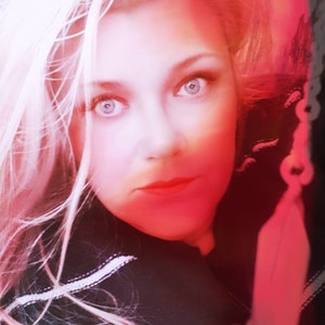 May include: A woman with long blonde hair and bright blue eyes is looking directly at the camera. Her face is illuminated by a red light, and her lips are painted a bright red. She is wearing a black shirt with a white stripe.