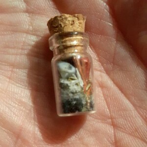 May include: A small glass bottle with a cork stopper filled with various small objects, including a white shell, a piece of red coral, and other small pieces of sea glass.