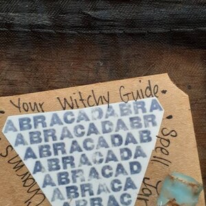 May include: A brown paper tag with the text "Your Witchy Guide" and "Spell Jars and Magick Charms". A white triangle sticker with the text "ABRACADABRA" repeated in a pattern is on the tag. A small glass jar with a blue lid and filled with small rocks and crystals is on the tag.