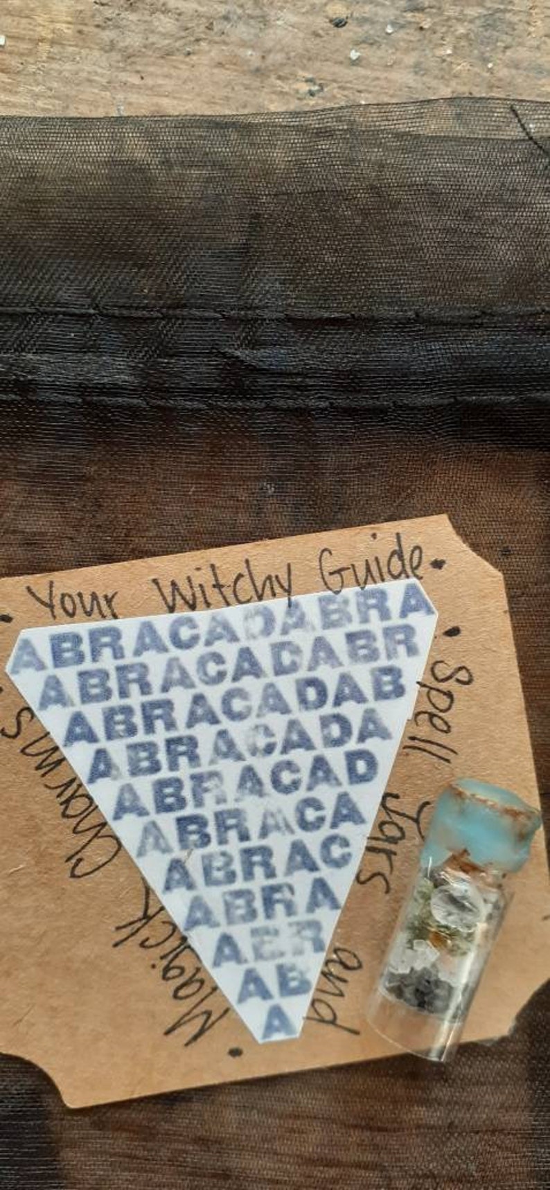 May include: A brown paper tag with the text "Your Witchy Guide" and "Spell Jars and Magick Charms". A white triangle sticker with the text "ABRACADABRA" repeated in a pattern is on the tag. A small glass jar with a blue lid and filled with small rocks and crystals is on the tag.