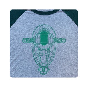 May include: A green and gray t-shirt with a green outline of a spaceship on the front. The spaceship is a top-down view of the Slave I from Star Wars.