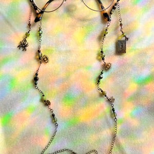 May include: Round spectacles with a decorative chain. The chain features green and black beads, small charms including a spider, a sun, and a small book, and a chain with an evil eye charm.