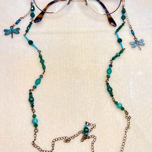 May include: A pair of round-framed spectacles with a gold-tone metal frame and tortoiseshell-patterned temple tips, attached to a beaded glasses chain featuring green and turquoise beads, bronze-tone metal links, and two small dragonfly charms.