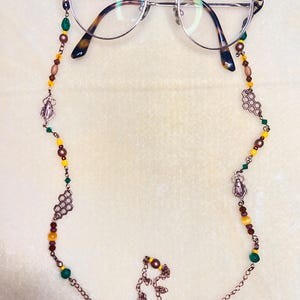 May include: A pair of round-framed spectacles attached to a beaded chain. The chain features copper-toned metal links, small green, yellow, and brown beads, and decorative charms shaped like insects and hexagonal honeycomb patterns.