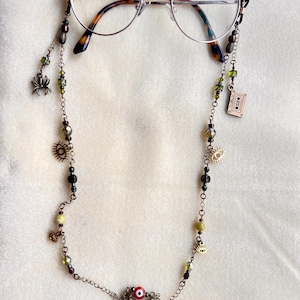 May include: Round-framed glasses with a tortoiseshell pattern on the temple tips, attached to a decorative metal chain. The chain features various beads in shades of green and brown, along with charms including a spider, a cassette tape, and eyes.