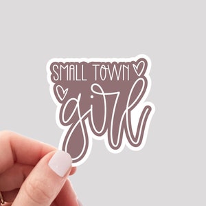 May include: A sticker with the words "SMALL TOWN GIRL" in a handwritten style. The text is in white with a mauve background and a white border. Two small hearts are above the word "TOWN" and one above the word "GIRL".