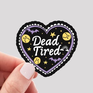 May include: A heart-shaped sticker with a black border and a purple interior. The sticker features the words "Dead Tired" in white, surrounded by stars, moons, bats, and sad face emojis. The text "WATERPROOF VINYL STICKER" is at the bottom.