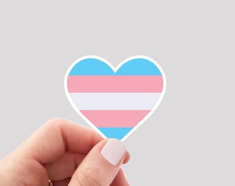 May include: A hand holding a heart-shaped sticker with the transgender pride flag colors: light blue, pink, white, and light blue.