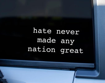 May include: A white vinyl decal applied to a dark tinted vehicle window. The decal features the text in a clean, sans-serif font that reads: hate never made any nation great. The lettering is arranged in three lines against the dark glass surface.