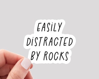 May include: White sticker with black text that reads "EASILY DISTRACTED BY ROCKS". A hand is holding the sticker.