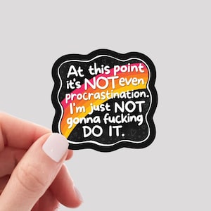 May include: A black, die-cut vinyl sticker with the text "At this point it's NOT even procrastination. I'm just NOT gonna fucking DO IT." The text is in white, pink, orange, and yellow. The sticker is held by a hand.