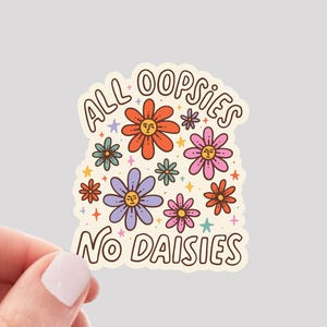 May include: A colorful sticker with the text "ALL OOPSIES NO DAISIES" in a retro font. The sticker features various flowers in orange, pink, purple, and teal. The words "WATERPROOF VINYL STICKER" are at the bottom.