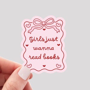 May include: A pink sticker with the text "Girls just wanna read books" in a decorative frame with hearts and a bow. The sticker is held by a hand and the words "WATERPROOF VINYL STICKER" are at the bottom.