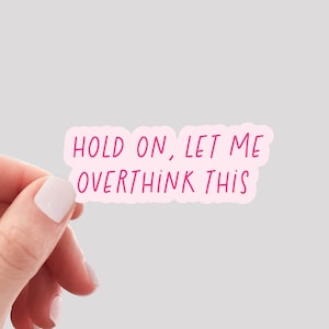 May include: A pink sticker with the phrase "HOLD ON, LET ME OVERTHINK THIS" in a handwritten style. The sticker is held by a hand with manicured nails against a light gray background.