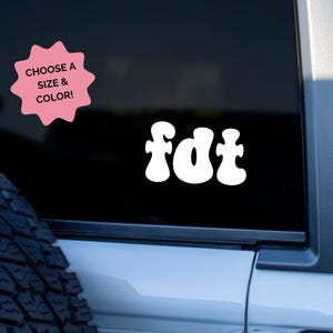 May include: White vinyl decal with the word "fdt" in a retro font. A pink starburst-shaped sticker in the upper left corner reads "Choose a Size & Color!" The decal is displayed on a black background.