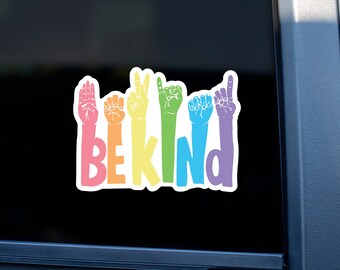 May include: A rainbow-colored sticker with the word "BE KIND" spelled out in sign language. The sticker is on a black car window.