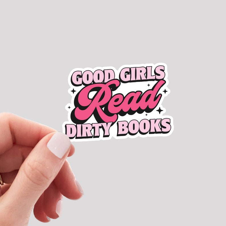 May include: A sticker with the text "GOOD GIRLS Read DIRTY BOOKS" in pink and black, with a white border. The word "Read" is in a cursive font. The sticker is held by a hand against a light gray background.
