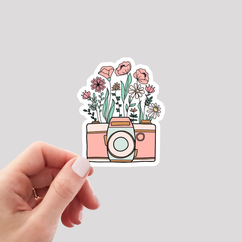May include: A pink and white camera with a floral design. The camera has a lens and a shutter button. The flowers are pink, white, and green.