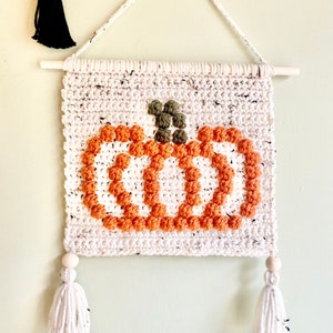 K&ouml;nnte beinhalten: Ein wei&szlig;er geh&auml;kelter Wandbehang mit einem braunen und orangefarbenen K&uuml;rbisdesign. Der K&uuml;rbis besteht aus Garnpompons und hat einen gr&uuml;nen Stiel. Der Behang hat zwei wei&szlig;e Quasten mit Holzperlen oben.