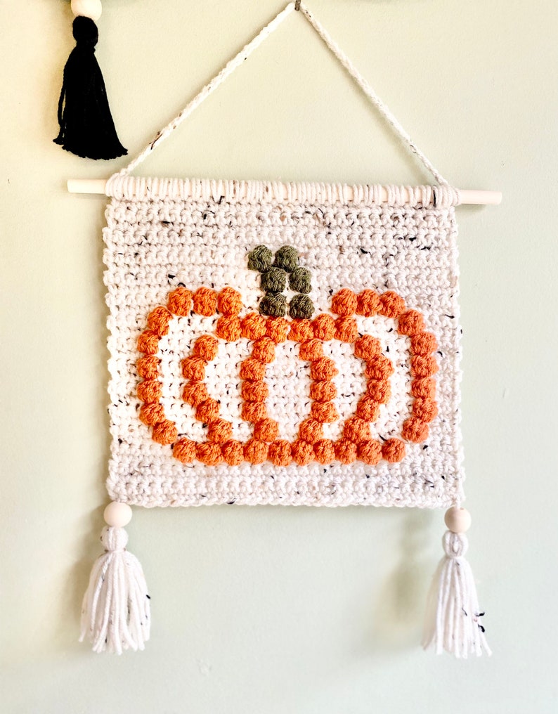 K&ouml;nnte beinhalten: Ein wei&szlig;er geh&auml;kelter Wandbehang mit einem braunen und orangefarbenen K&uuml;rbisdesign. Der K&uuml;rbis besteht aus Garnpompons und hat einen gr&uuml;nen Stiel. Der Behang hat zwei wei&szlig;e Quasten mit Holzperlen oben.