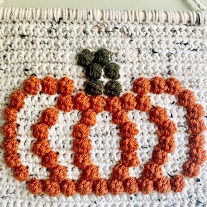 K&ouml;nnte beinhalten: Ein geh&auml;kelter K&uuml;rbis-Wandbehang mit wei&szlig;em Hintergrund und orangefarbenem und gr&uuml;nem Garn. Der K&uuml;rbis besteht aus einer Reihe kleiner, runder Bommeln.