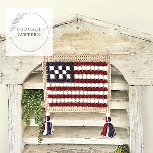 May include: A crocheted American flag wall hanging with red, white, and blue pom-poms. The flag is hanging on a white wooden wall with a wooden shelf below it. The flag has tassels on the bottom.