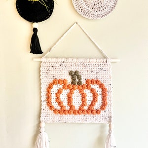 K&ouml;nnte beinhalten: Ein wei&szlig;er geh&auml;kelter Wandbehang mit einem braunen und orangefarbenen K&uuml;rbisdesign. Der K&uuml;rbis ist aus strukturiertem Garn gefertigt und hat einen gr&uuml;nen Stiel. Der Wandbehang hat zwei wei&szlig;e Quasten mit Holzperlen am unteren Ende.