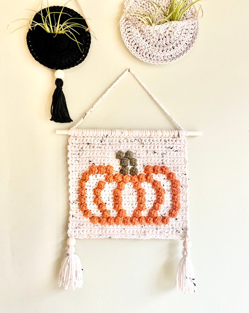 K&ouml;nnte beinhalten: Ein wei&szlig;er geh&auml;kelter Wandbehang mit einem braunen und orangefarbenen K&uuml;rbisdesign. Der K&uuml;rbis ist aus strukturiertem Garn gefertigt und hat einen gr&uuml;nen Stiel. Der Wandbehang hat zwei wei&szlig;e Quasten mit Holzperlen am unteren Ende.