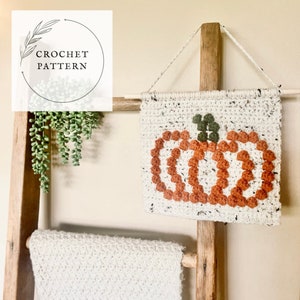 K&ouml;nnte beinhalten: Ein geh&auml;kelter K&uuml;rbis-Wandbehang mit wei&szlig;em Hintergrund und orangefarbenem und gr&uuml;nem Garn. Der Behang befindet sich auf einer Holzleiter mit einer wei&szlig;en H&auml;keldecke.