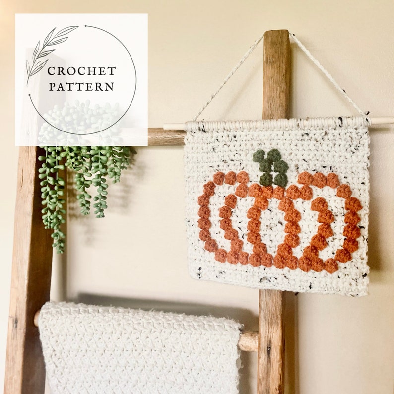 K&ouml;nnte beinhalten: Ein geh&auml;kelter K&uuml;rbis-Wandbehang mit wei&szlig;em Hintergrund und orangefarbenem und gr&uuml;nem Garn. Der Behang befindet sich auf einer Holzleiter mit einer wei&szlig;en H&auml;keldecke.