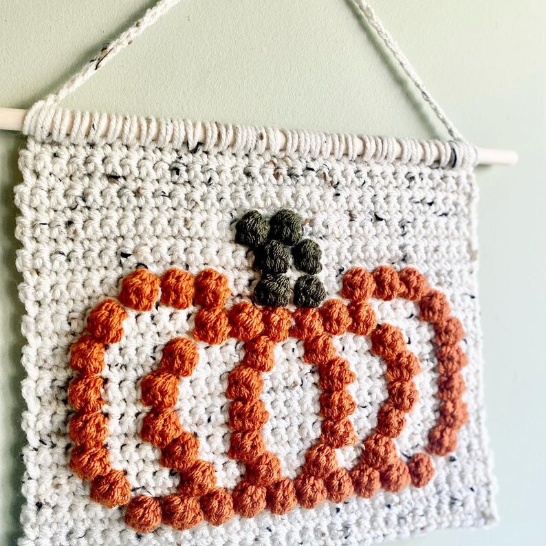 K&ouml;nnte beinhalten: Ein geh&auml;kelter Wandbehang mit einem K&uuml;rbisdesign. Der K&uuml;rbis ist aus orangefarbenem und braunem Garn gefertigt und hat einen gr&uuml;nen Stiel. Der Hintergrund ist ein wei&szlig;er geh&auml;kelter Stoff.
