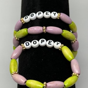 May include: Two bracelets with the word "DOPEY" spelled out in white beads with black lettering. The bracelets are made of wooden beads in shades of green and purple.