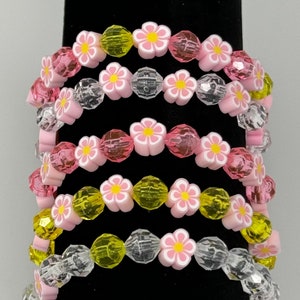 May include: Five pink and white flower bead bracelets with clear, pink, and yellow beads. The bracelets are stacked on a black surface.