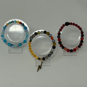 May include: Three beaded bracelets on clear stands. One bracelet is blue with the word "SPELL" in colorful letters. Another has the word "FALLOUT" in yellow beads and a lightning bolt charm. The third is red with the word "NEW" in red letters.