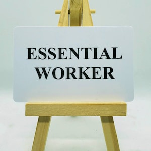 May include: A white card with the text "ESSENTIAL WORKER" displayed on a small wooden easel.