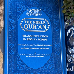 May include: A blue book titled "The Noble Qur'an" with gold lettering and decorative borders. The book contains transliteration in Roman script and English translation. The cover also features Arabic text.