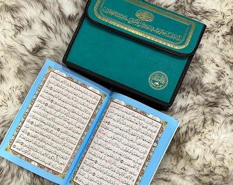 May include: A teal blue leather-bound book with gold lettering and a decorative gold circle on the cover. The book is open to a page with Arabic text. The book is sitting on a fluffy white surface.