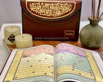 May include: An open book with Arabic script, featuring colorful text and decorative borders. A brown leather case with gold calligraphy sits behind the book. Two candles and a small vase with reeds are also visible.