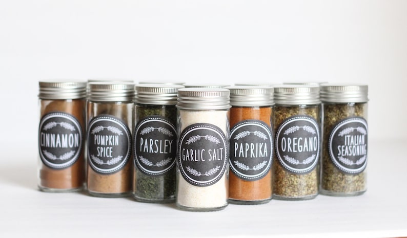 K&ouml;nnte beinhalten: Eine Reihe von acht Glas-Gew&uuml;rzgl&auml;sern mit schwarzen Etiketten und wei&szlig;em Text. Die Etiketten beinhalten "Zimt", "K&uuml;rbisgew&uuml;rz", "Petersilie", "Knoblauchsalz", "Paprika", "Oregano" und "Italienische W&uuml;rze".