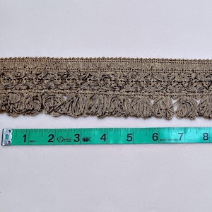 May include: A decorative trim with a metallic gold color, featuring a detailed floral pattern and fringe. The trim is measured with a green tape measure, showing a length of approximately 14 inches.