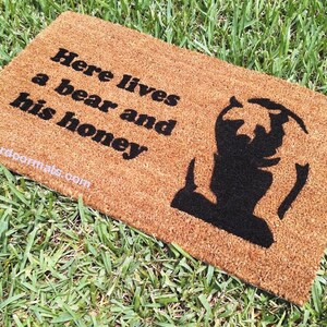 May include: A brown coir doormat with the text "Here lives a bear and his honey" in black lettering. A black silhouette of a bear is on the right side of the mat.