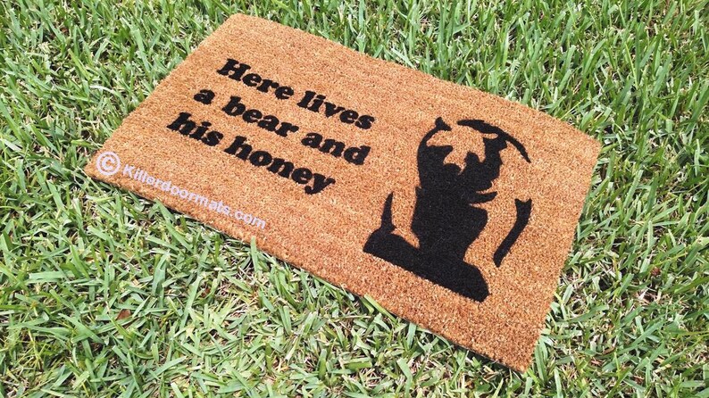 May include: A brown coir doormat with the text "Here lives a bear and his honey" in black lettering. A black silhouette of a bear is on the right side of the mat.